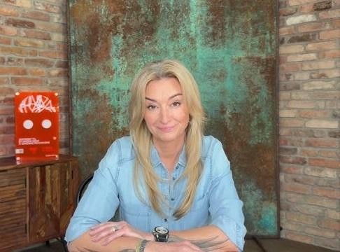 Martyna Wojciechowska, dziennikarka i podróżniczka, z mikrofonem, na tle mapy świata i sprzętu podróżniczego.