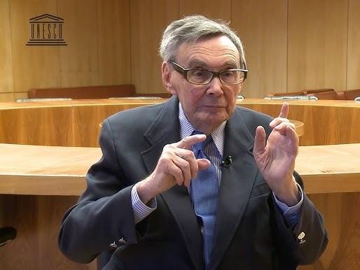 Marian Turski wygłaszający przemówienie podczas obchodów rocznicy wyzwolenia Auschwitz, z tłem obozowych baraków.