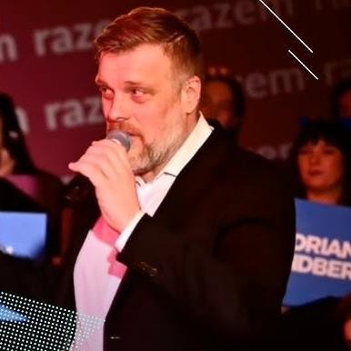 Adrian Zandberg, polityk Partii Razem, w zbliżeniu, z przyjaznym wyrazem twarzy, na tle niewyraźnego otoczenia.