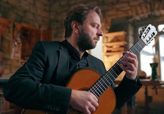 Marcin Dylla w zamyślonej pozie z gitarą, przygotowujący się do występu lub ćwiczący, w tle rozmyte wnętrze sali.