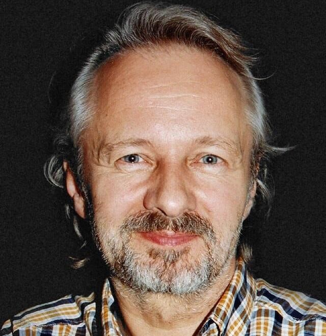 Krzysztof Stroiński w zamyśleniu, w swobodnym stroju, w tle rozmyte elementy planu filmowego lub teatralnego, oddające atmosferę skupienia na pracy aktorskiej.