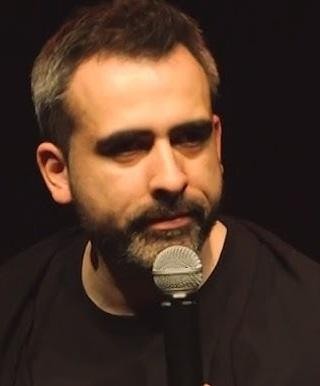 Abelard Giza podczas występu stand-upowego, z ekspresyjną mimiką, trzymający mikrofon.