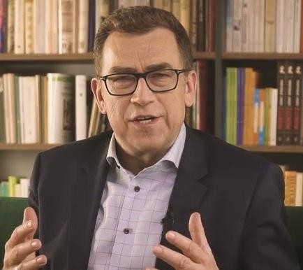 Maciej Orłoś podczas szkolenia z wystąpień publicznych, gestykulujący, z zaangażowanym wyrazem twarzy, w tle rozmyta sala konferencyjna.