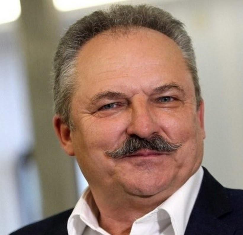Marek Jakubiak w eleganckim garniturze, w biurze, z zamyślonym wyrazem twarzy, w tle subtelne elementy nawiązujące do jego działalności.